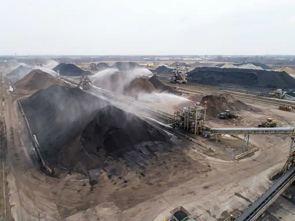 Luchtfoto van een droge bulkterminal met grote steenkoolopslagpiles, stofonderdrukking en industriële transportbanden op de achtergrond.