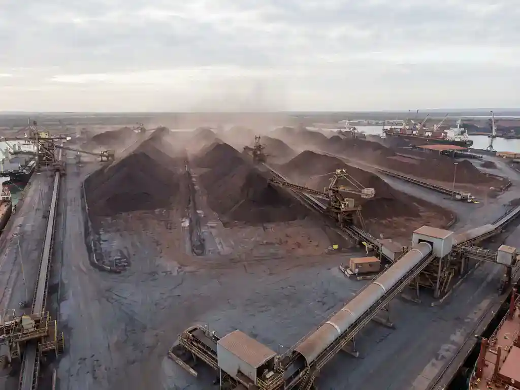 Luchtfoto van een groot ijzerertsoverslagterminal met roodbruine ertshopen, transportbanden en stofnevel in diffuus daglicht.